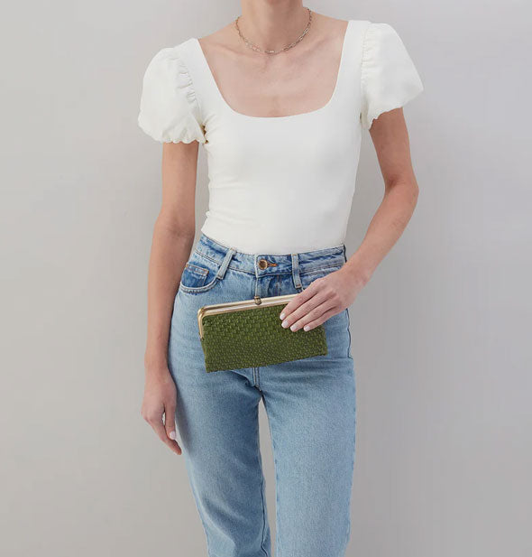 Model in jeans and a white blouse stands holding a green woven leather wallet with gold-toned frame hardware
