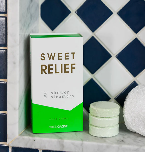 Sweet Relief shower steamers box and three light green steamer discs rest on a marble bath ledge with a rolled-up washcloth in front of black and white tile
