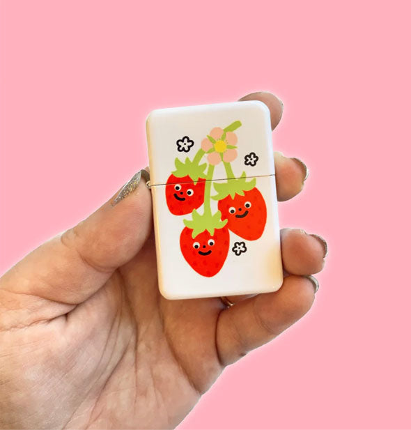 Model's hand holds a rectangular white flip-top lighter with illustration of three smiling red strawberries on a green vine with yellow flower flanked by small flower accents