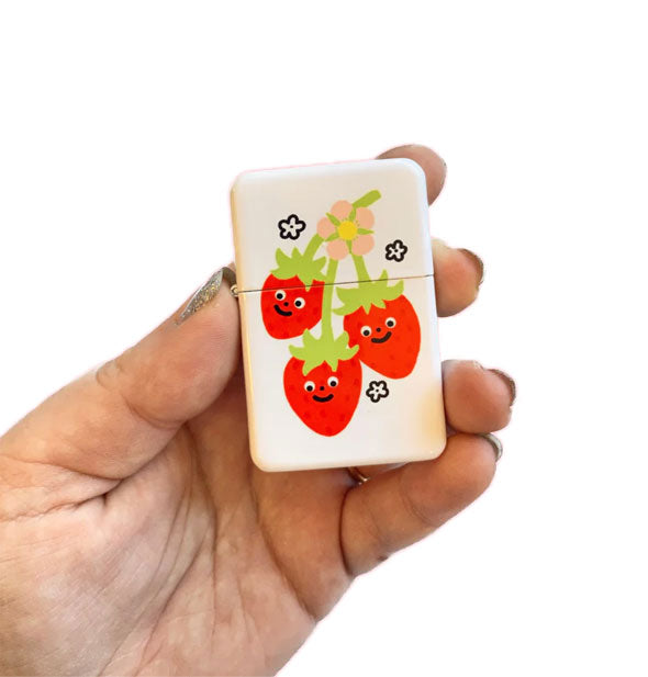 Model's hand holds a rectangular white flip-top lighter with illustration of three smiling red strawberries on a green vine with yellow flower flanked by small flower accents