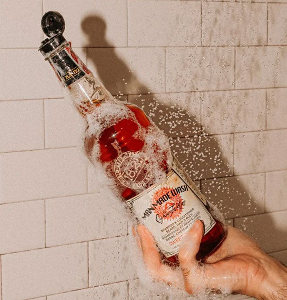 Model's hand holds a bottle of 18.21 Mad Made Wash covered in suds under a shower stream against a white tiled backdrop