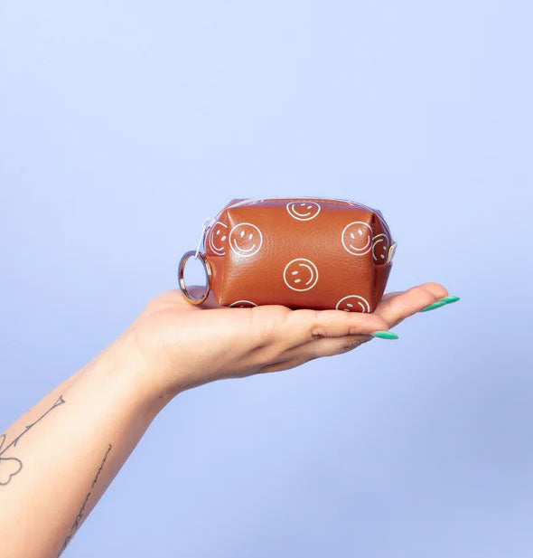 Model's hand holds a brown vegan leather smiley face print keyring pouch up in front of a blue backdrop