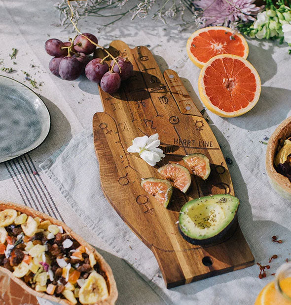 Wooden hand board is staged with fruits, avocado, and flowers on a tabletop