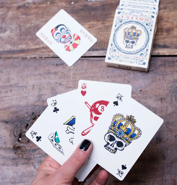 Model's hand holds a sample spread of Tattoo Playing Cards while the Joker and deck box rest on a rustic wooden surface behind