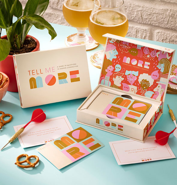 Components of the Tell Me More connection game are staged with beer glasses, pretzels, darts, and a houseplant