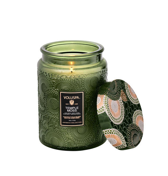 Green embossed glass Voluspa Temple Moss candle with matching green, gold, and cream floral patterned metal lid removed and propped up against it