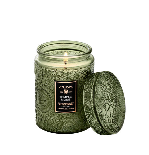 Lit embossed green glass Voluspa Temple Moss candle with lid removed and propped against it