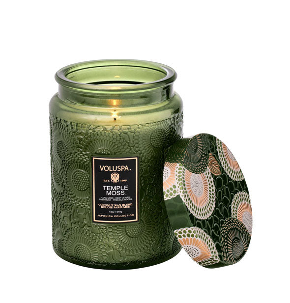Green embossed glass Voluspa Temple Moss candle with matching green, gold, and cream floral patterned metal lid removed and propped up against it