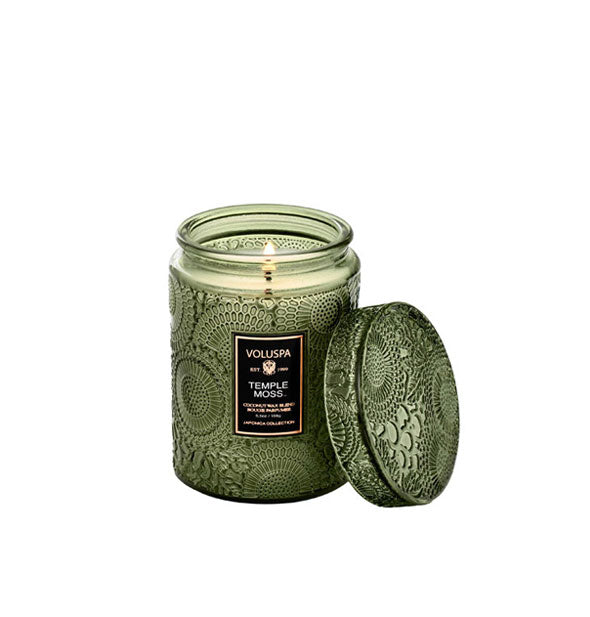 Lit embossed green glass Voluspa Temple Moss candle with lid removed and propped against it