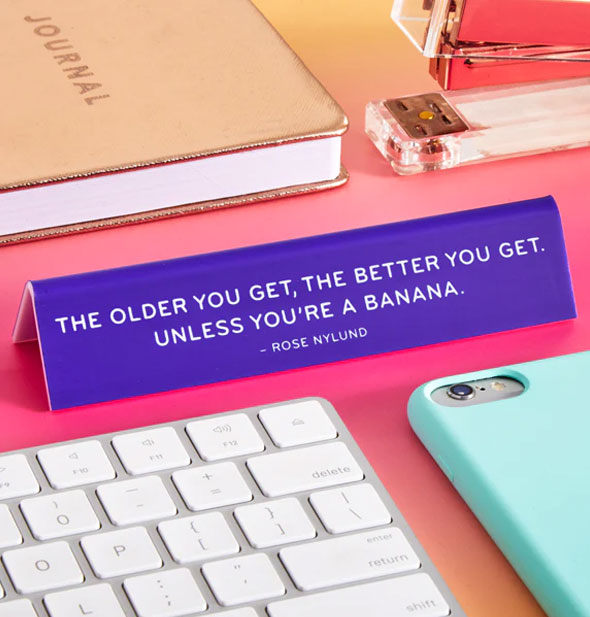 Purple Rose Nylund banana quote desk sign staged with computer keyboard, smartphone, gold journal, and stapler on a pink surface