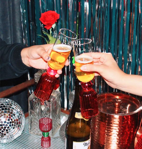 Models' hands hold two Tipsy Turvy Bar Glasses in a toast against a festive-looking backdrop
