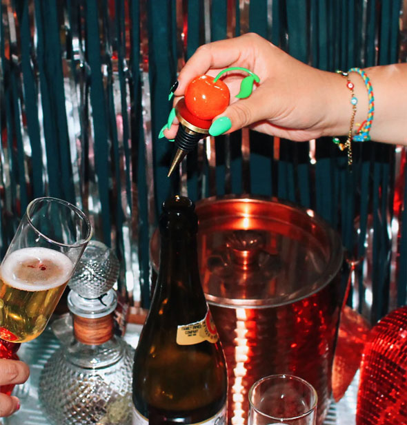 Model's hand removes a cherry stopper from a bottle of champagne against a festive-looking backdrop
