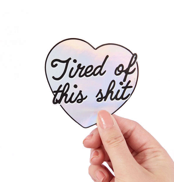 Model's hand holds a holographic heart-shaped sticker that says, "Tired of this shit" in black script