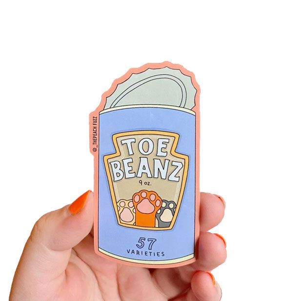 Model's hand holds a sticker shaped like a can with label that says, "Toe Beanz" with image of cat paws