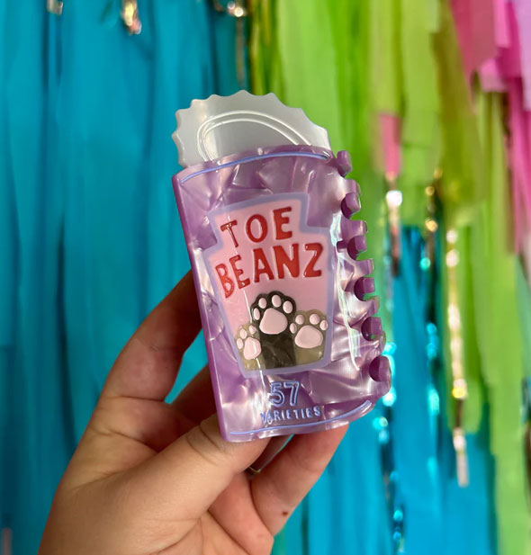 Model's hand holds a Toe Beanz claw clip with cat paws design in front of a colorful streamer backdrop