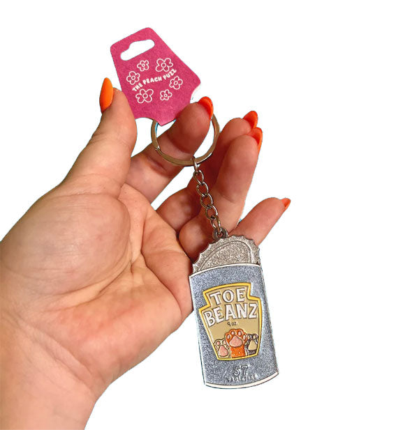 Model's hand holds a glittery Toe Beanz keychain designed as an opened food can with cat paws on the label