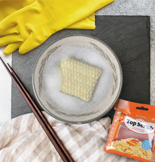 Ramen noodle-shaped dish sponge rests on a dinner plate next to chopsticks and yellow kitchen gloves