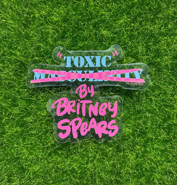 Clear sticker with pink and blue text that reads, "'Toxic' Masculinity [crossed out] by Britney Spears" rests on green astroturf