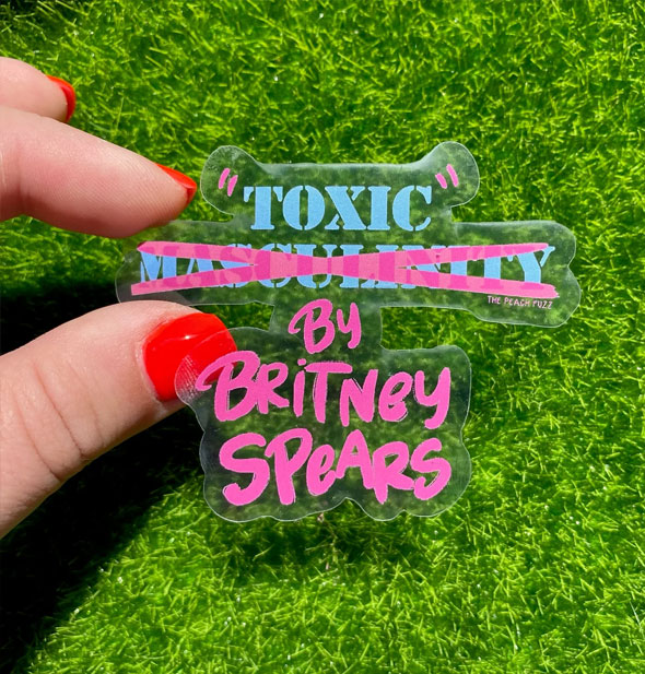 Model's hand holds a "Toxic" by Britney Spears sticker slightly above green astroturf