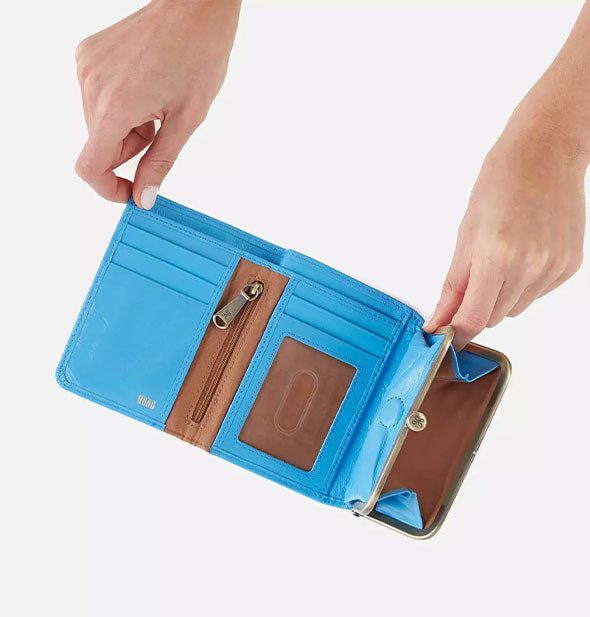 Model's hands hold a blue leather wallet open to show slots and pockets inside with brown lining detail and gold hardware