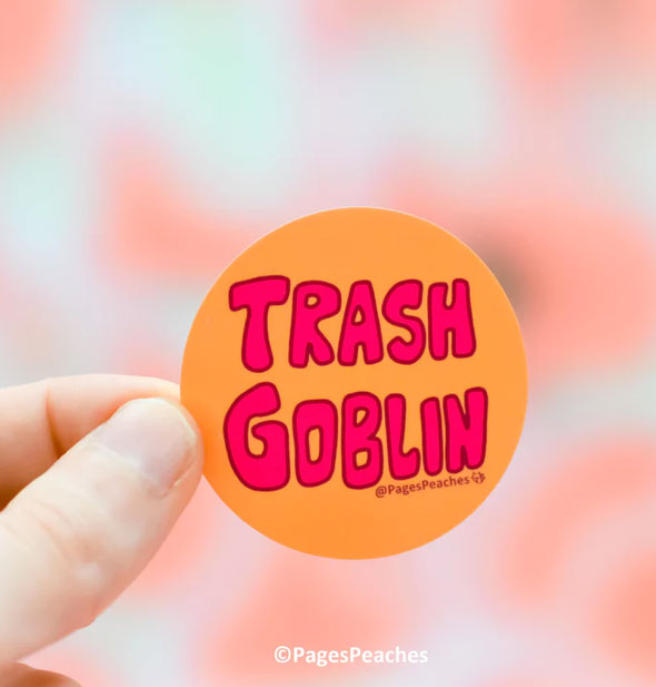 Model's hand holds a round orange sticker that says, "Trash Goblin" in large pink lettering
