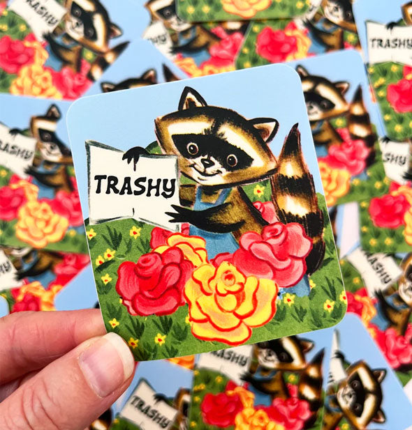 Model's hand holds a Trashy raccoon sticker in front of a pile of others like it