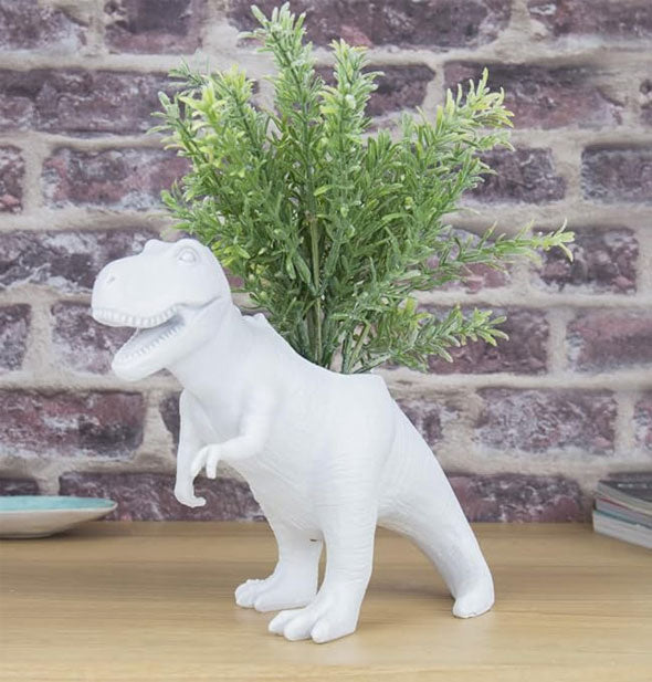 T-Rex planter staged with fern-like greenery sticking out of its back against a brick backdrop