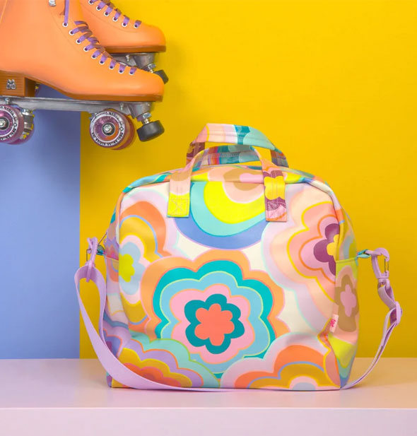A Trippy Dippy Floral Skate Bag rests on a pink surface against a mustard and periwinkle backdrop near a pair of suspended orange roller skates