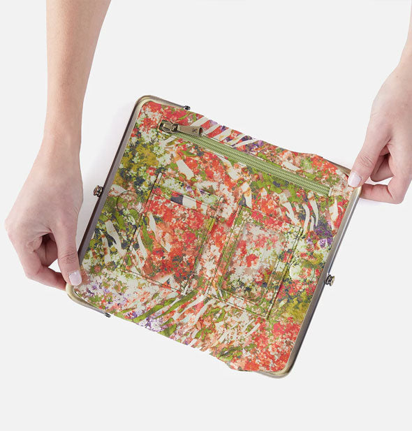 Model's hands hold open a multicolored tropical floral print wallet with brass hardware, interior slip pickets, and green zipper pocket