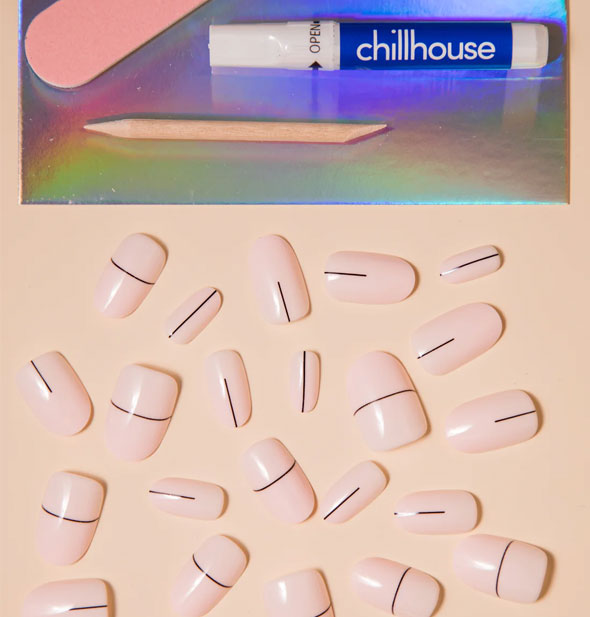 A smattering of pale whitish-pink press-on nails with single thin, black lines on each next to Chillhouse glue tube, pink nail file, and wooden cuticle pusher on an iridescent tray