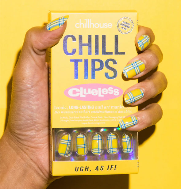 Model's hand wears a set of yellow and blue plaid Chillhouse Chill Tips Clueless edition press-on nails and holds a yellow box of the same design, called Ugh, As If!