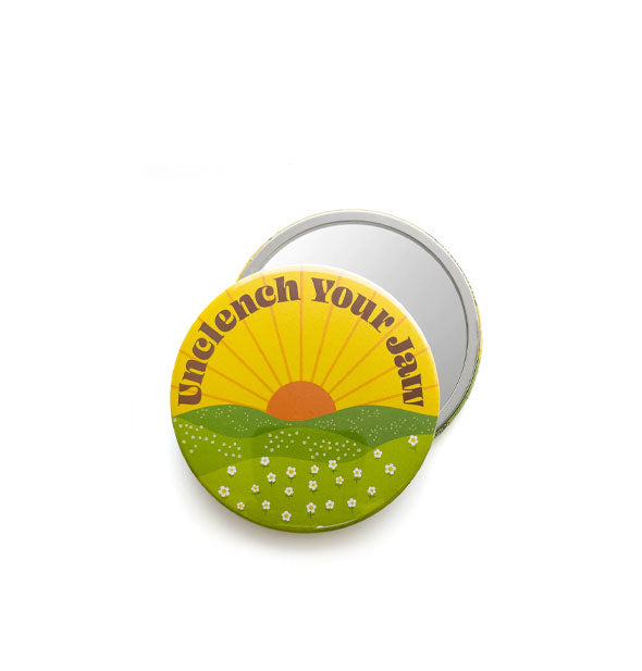 Front and back views of a round pocket mirror that features artwork of a sun rising over rolling hills covered in white flowers and the words, "Unclench Your Jaw" printed at the top