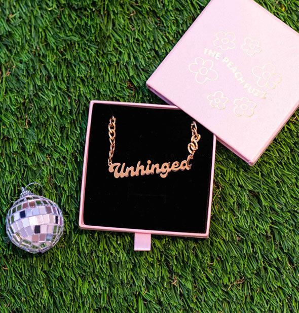 Gold "Unhinged" curb chain nameplate necklace in a slide-open purple box with black felt lining rets on green astroturf next to a mini disco ball