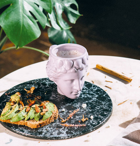Purple Venus head mug filled with a foamy beverage rests on a round black marble tray next to a piece of half-eaten avocado toast