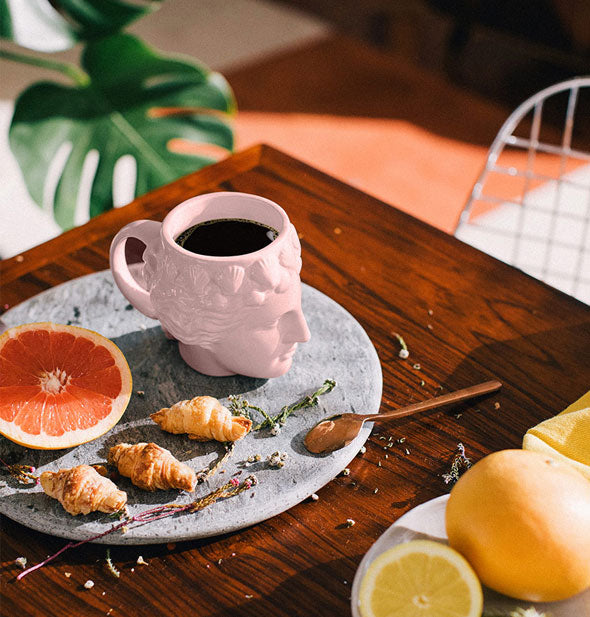 Pink Venus head mug with black coffee inside is staged on a stone slab on a wooden tabletop with grapefruit, mini croissants, and other brunch accouterments