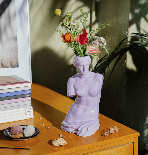 Purple Venus de Milo vase on wooden tabletop with stones, dried flowers, and books holds a small bouquet of flowers and greens