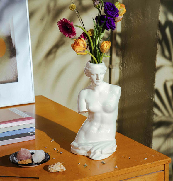 White Venus de Milo vase on top of a wooden tabletop with stones, dried flowers, and books holds a small bouquet of colorful flowers