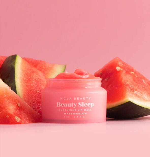 Opened pot of Watermelon NCLA Beauty brand Beauty Sleep Overnight Lip Mask staged with pieces of cut watermelon against a pink backdrop