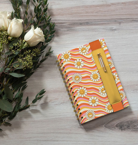 Waves of Melody notebook with pen sleeve on a wooden surface next to a bouquet of white roses