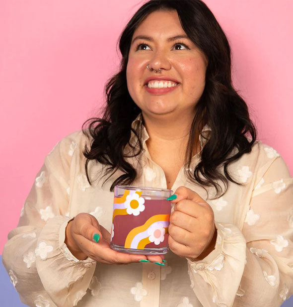 Smiling model holds a Wavy Daisy glass mug filled with a purplish beverage with both hands in front of a pink and blue backdrop