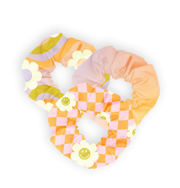 Set of three hair scrunchies: one with colorful stripes and white daisies, one in a pastel ombre, and one with pink and orange checker print accented with smiley face daisies