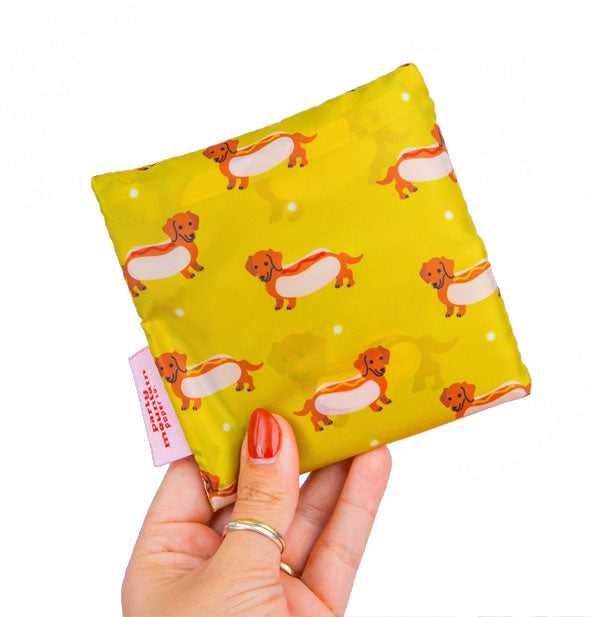 Model's hand holds a folded-up mustard yellow tote bag with print of Dachshunds in hot dog buns