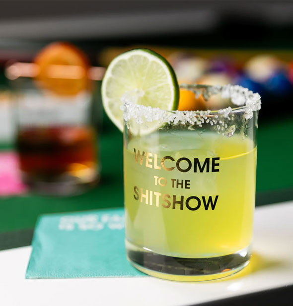 Welcome to the Shitshow rocks glass resting on a teal napkin is filled with a lime-colored beverage and garnished with lime slice and salted rim