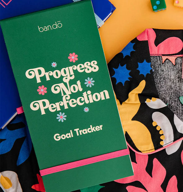 Green Progress Not Perfection Goal Tracker pad with elastic pink band rests on a colorful surface
