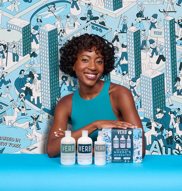 Smiling model poses with the contents of a Verb Where's Hydrate? Holiday Kit against a matching backdrop of blue "Raised in New York" illustrated design