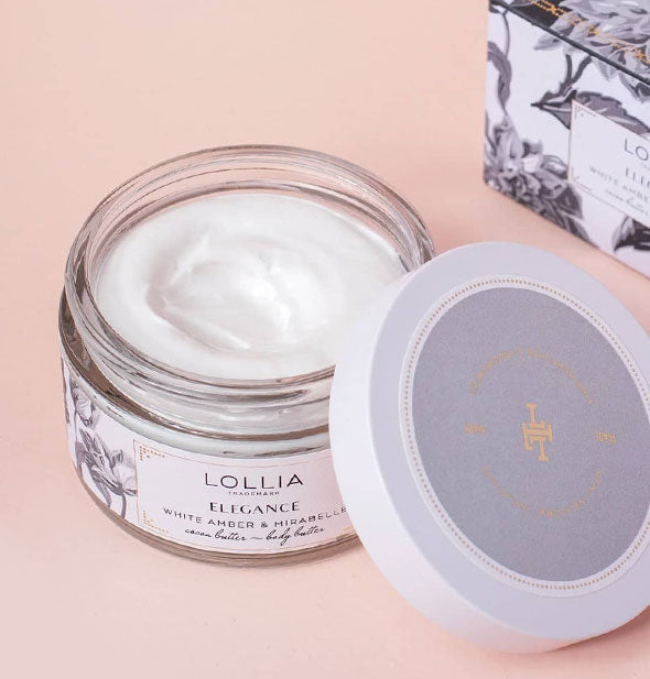 Opened pot of Lollia Elegance Body Butter with lid set to the side and box behind reveals creamy white product inside
