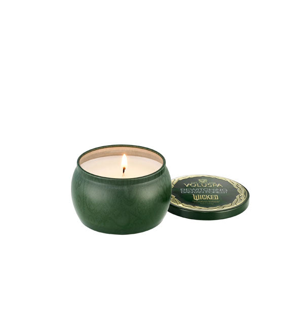 Lit Voluspa Bewitched Banyan Forest Wicket-themed candle inside a green tin with matching gold-framed lid removed and set to the side