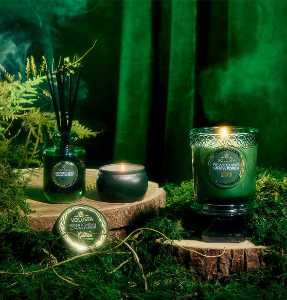 Wicked-themed green Voluspa Bewitching Banyan Forest candles and reed diffuser staged with wood, ferns, and moss against a misty green velvet backdrop