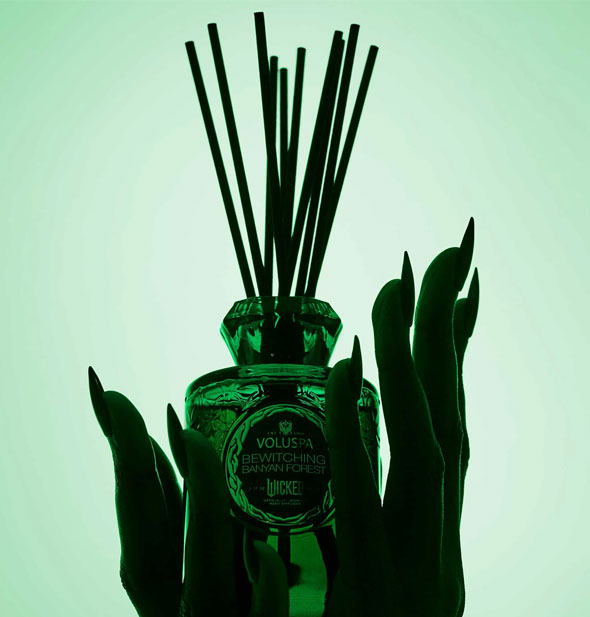 Silhouetted hands with long fingernails hold an embossed green glass Wicked Bewitching Banyan Forest Voluspa reed diffuser