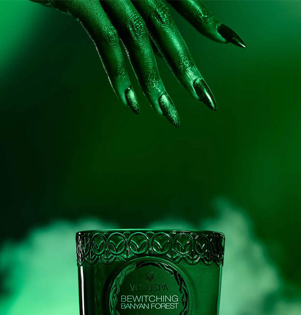 A model's fingertips with long, shimmering fingernails hover above an embossed Voluspa Bewitching Banyan Forest candle jar set against a misty green backdrop